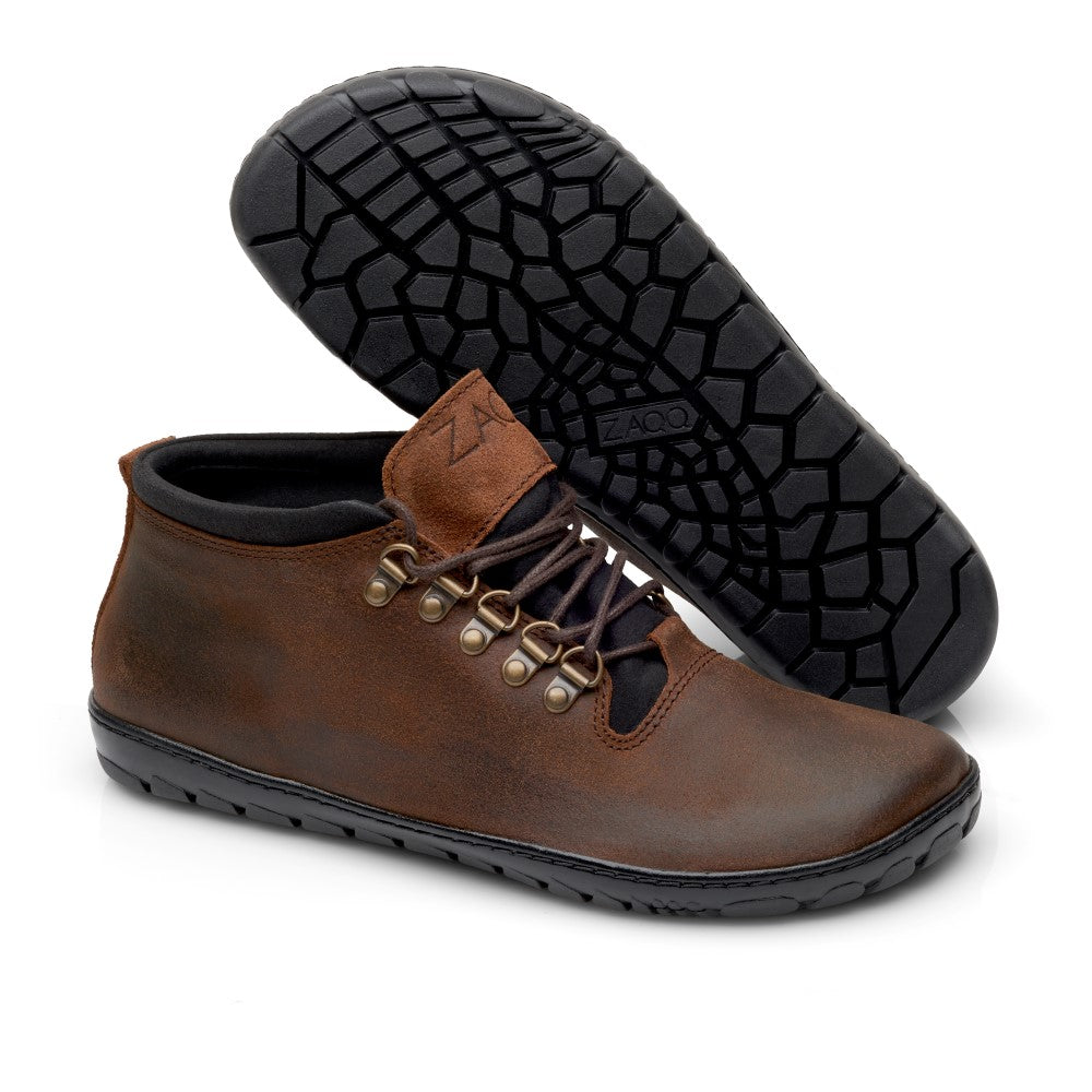 Ein Paar wasserdichte Lederstiefeletten EXPEQ Mid Brown von ZAQQ mit schwarzer Gummisohle und Metallösen. Ein Stiefel steht aufrecht, während der andere auf der Seite liegt, um das strukturierte Muster der Barfußsohle zu präsentieren.