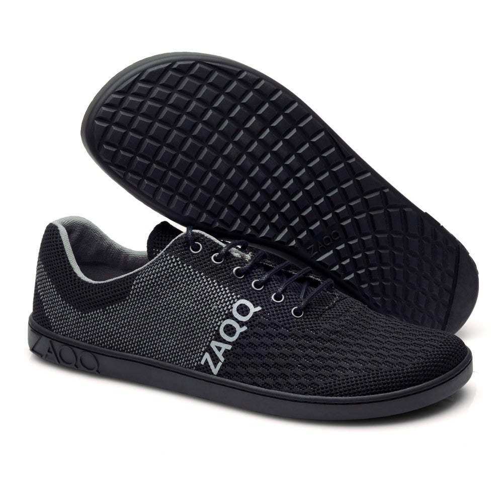 Ausgestellt werden die veganen Sportschuhe QNIT Black von ZAQQ aus Strick. Ein Schuh steht aufrecht, um die Seite und die Schnürsenkel zu präsentieren, während der andere flach liegt und das gekreuzte Profilmuster auf seiner strukturierten Sohle hervorhebt. Der Markenname ZAQQ ist auf diesen stilvollen Kreationen deutlich sichtbar.