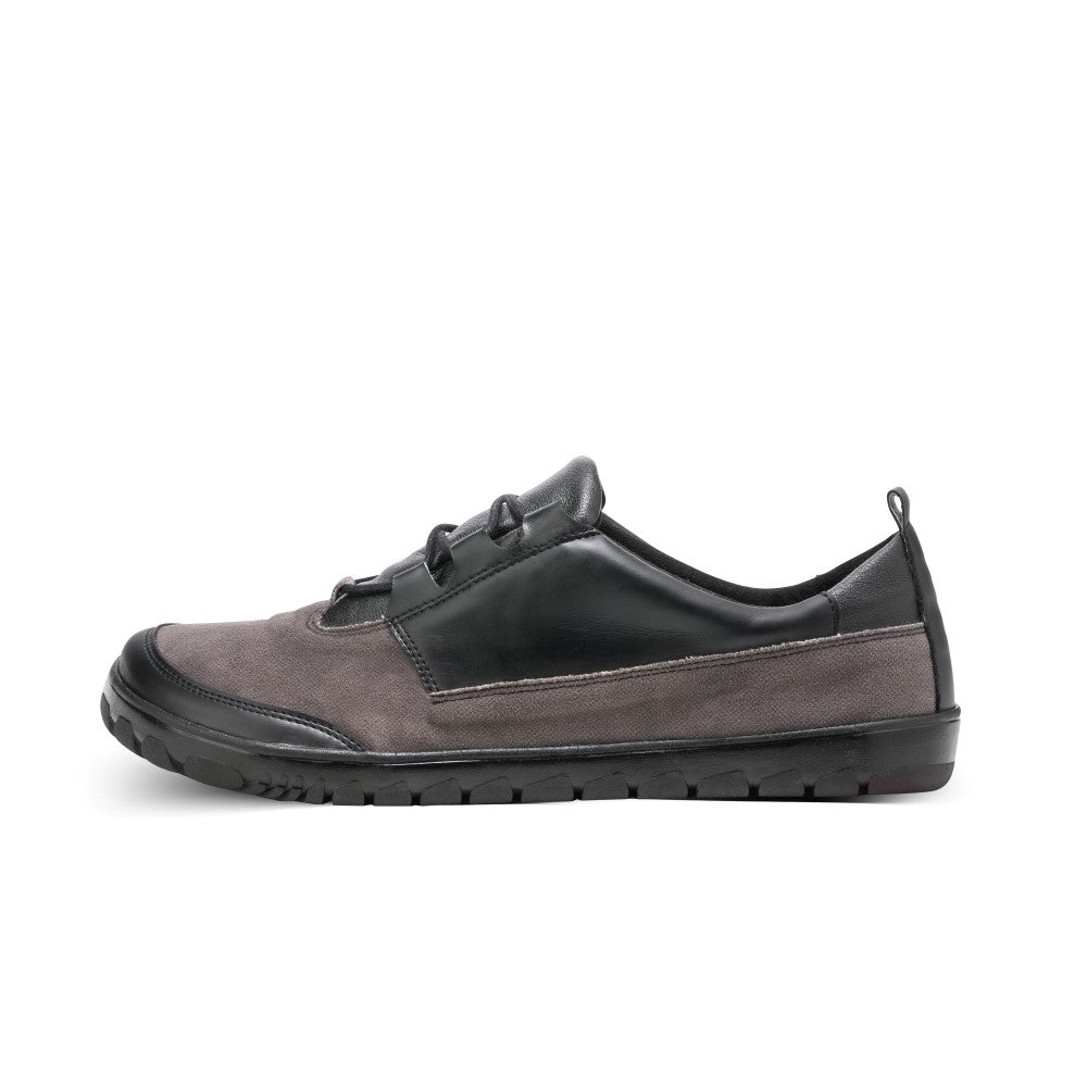 Der QUEST Low Black Waterproof-Schuh von ZAQQ besticht durch ein elegantes Design in Seitenansicht, mit einer grauen Wildlederfront, einem schwarzen Lederakzent an der Seite und einer schwarzen Sohle. Dieses minimalistische Schuhwerk ist wasserdicht und somit ideal für jedes Abenteuer.