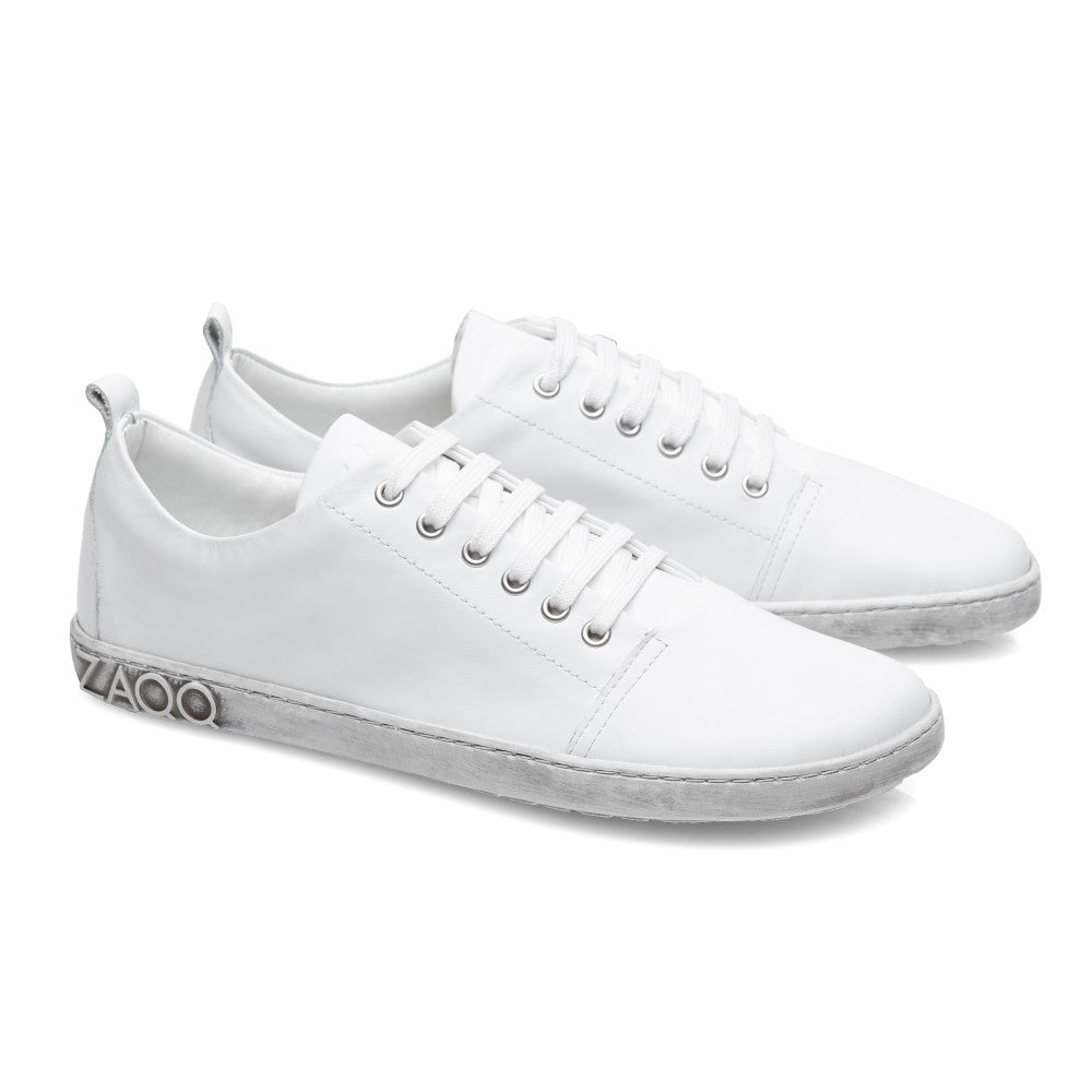 Der TAQQ Nappa White von ZAQQ ist ein weißer Sneaker aus hochwertigem Nappaleder mit Schnürsenkeln und niedrigem Schaft. Beide Schuhe haben graue Kautschuksohlen mit dem Aufdruck „Ziloco“ in stilisierter Schrift auf der Ferse.